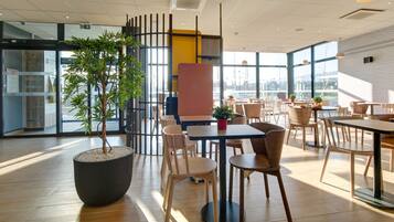 A modern indoor dining area with wooden tables and chairs, large windows, and a potted plant.