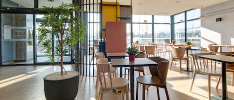 A modern indoor dining area with wooden tables and chairs, large windows, and a potted plant.