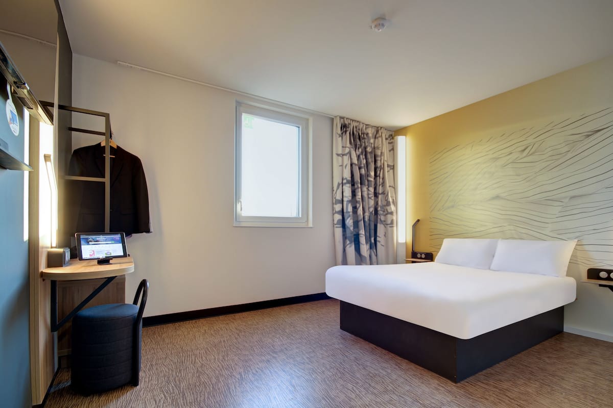 A modern hotel room with a bed, a desk with a computer, a chair, and a window with curtains.
