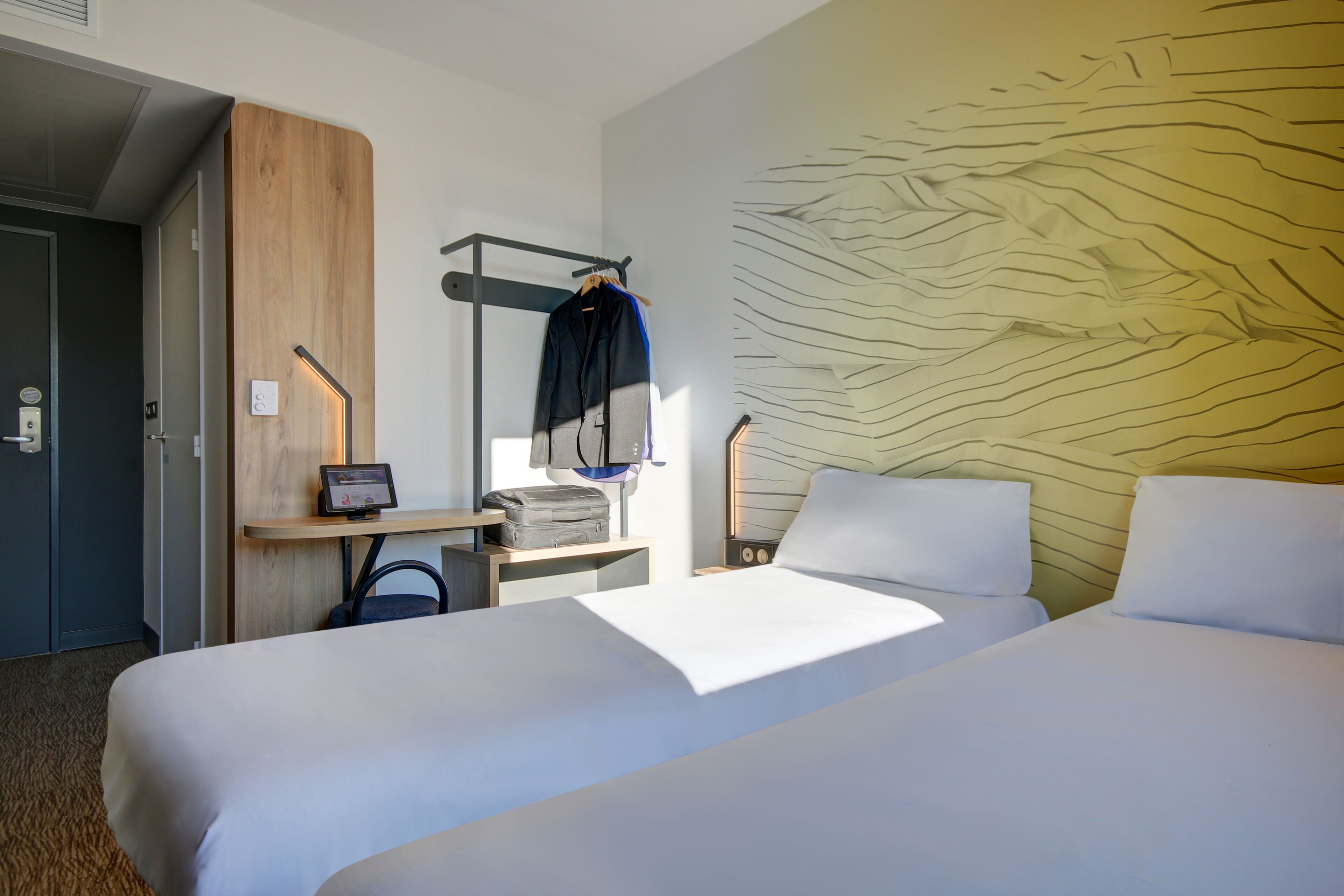 Ein Hotelzimmer mit zwei Betten, einem Schreibtisch mit Computer, einem Kleiderhaken mit Jacke und einer Wand mit strukturierter Gestaltung.