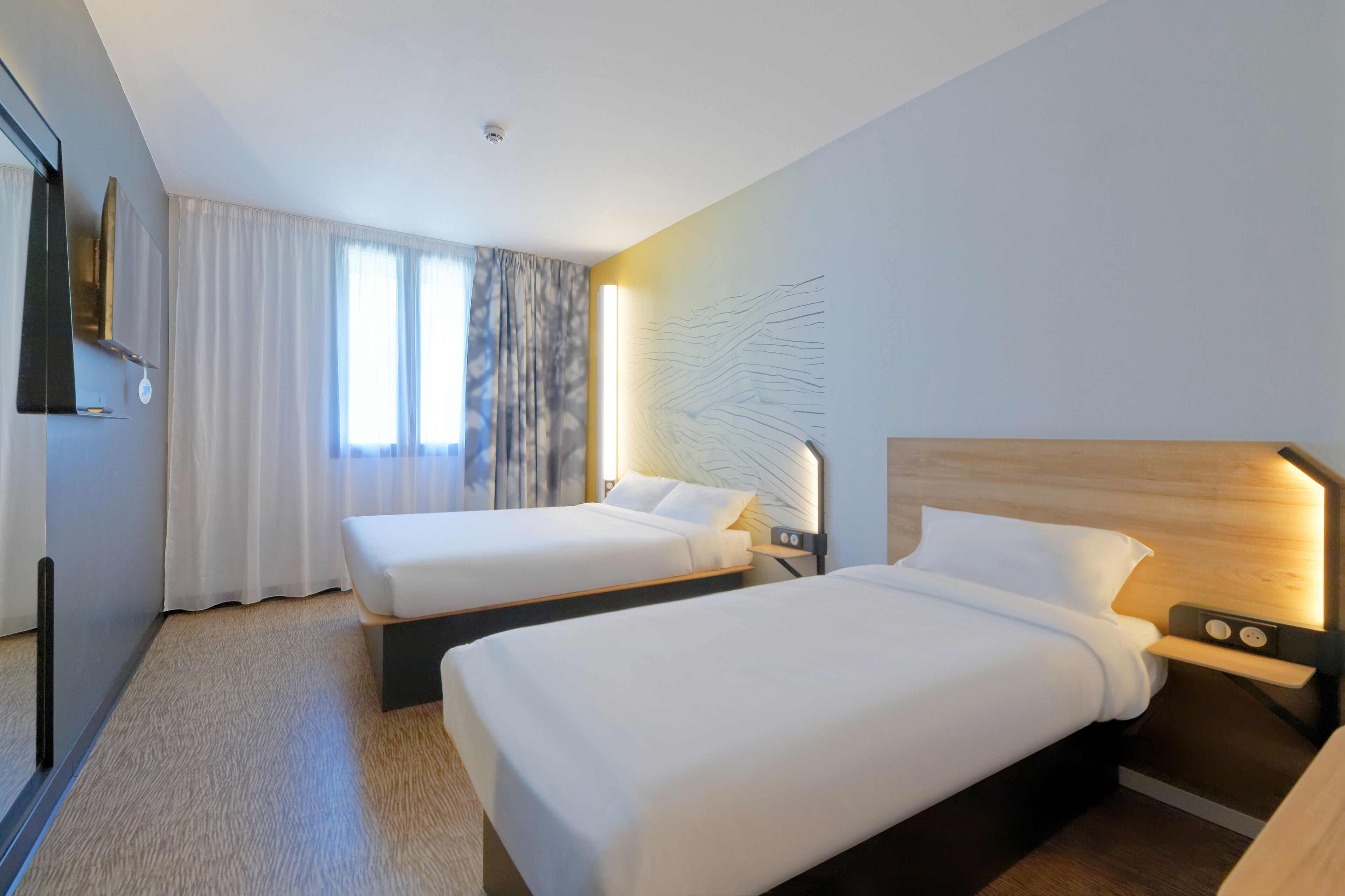 A hotel room with two beds, a large window with curtains, a wall-mounted TV, and a modern light fixture.
