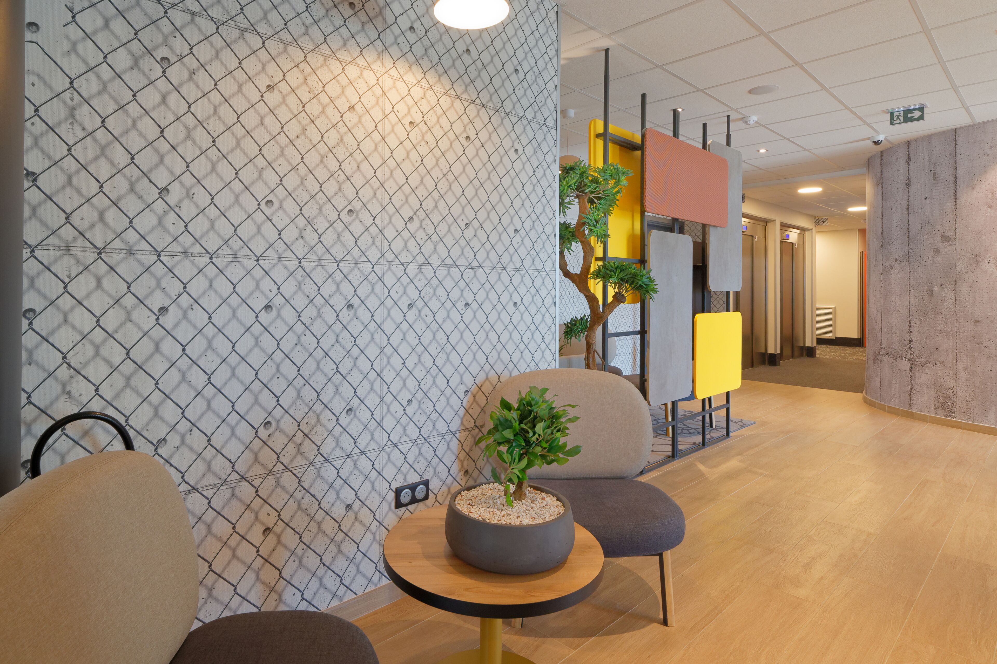 A modern interior with a chain-link partition, a round table with a potted plant, and a seating area with a yellow and orange accent wall.