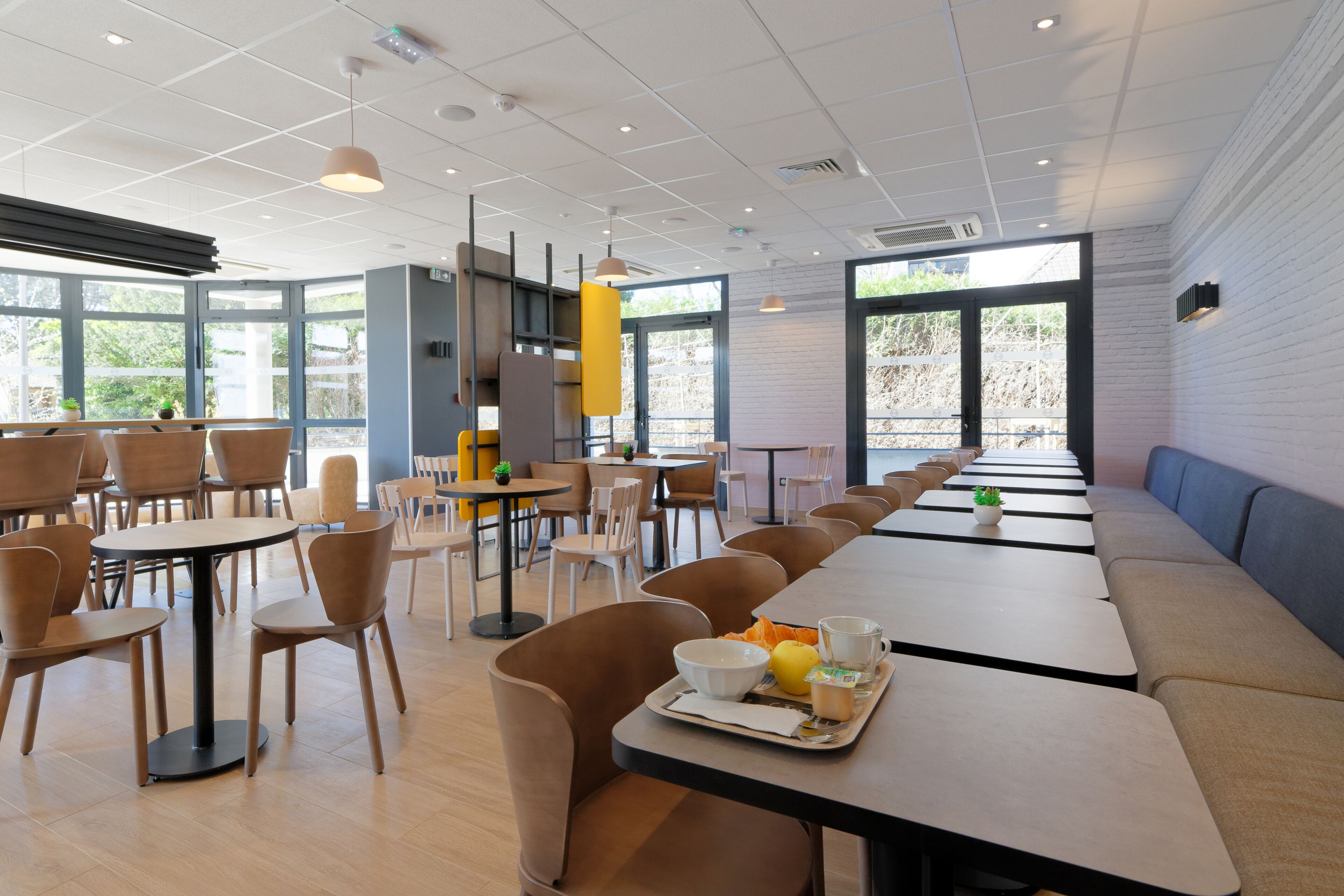Un área de comedor moderna con varias mesas y sillas, un banco largo, y una bandeja con comida y bebidas.