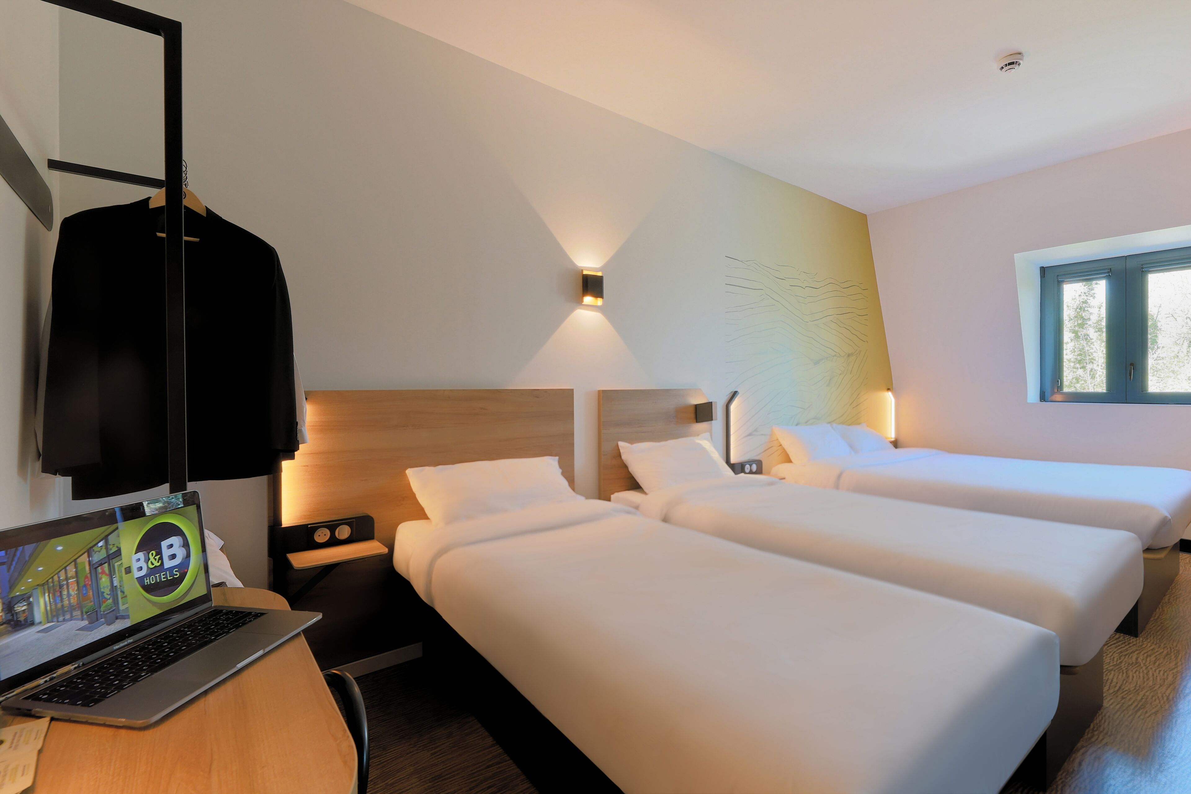 A hotel room with two single beds, a desk with a laptop, a nightstand, and a wall-mounted light fixture.