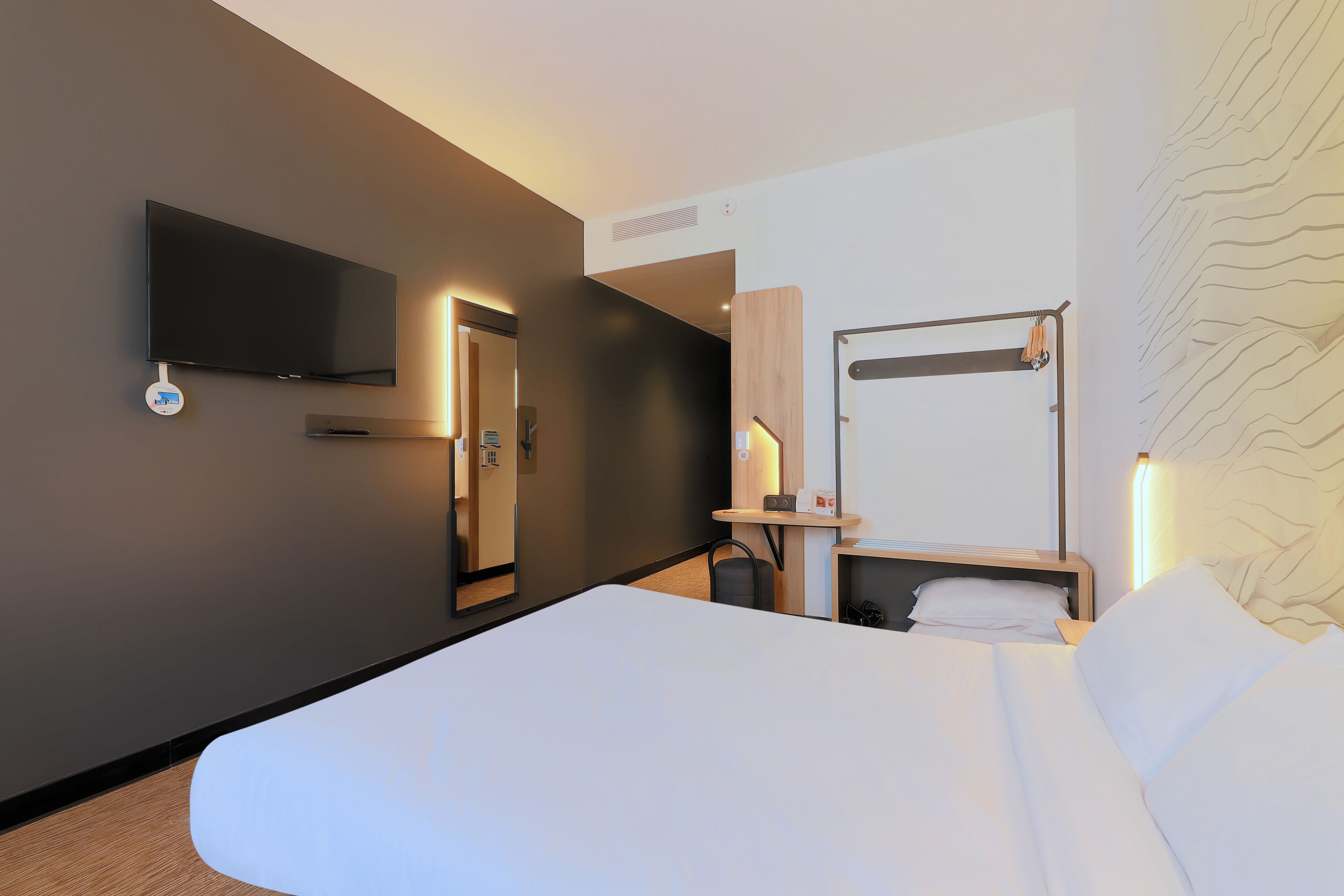 Une chambre d’hôtel moderne dotée d’un grand lit, d’un téléviseur à écran plat fixé au mur, d’une table de chevet avec une lampe et d’un petit bureau.