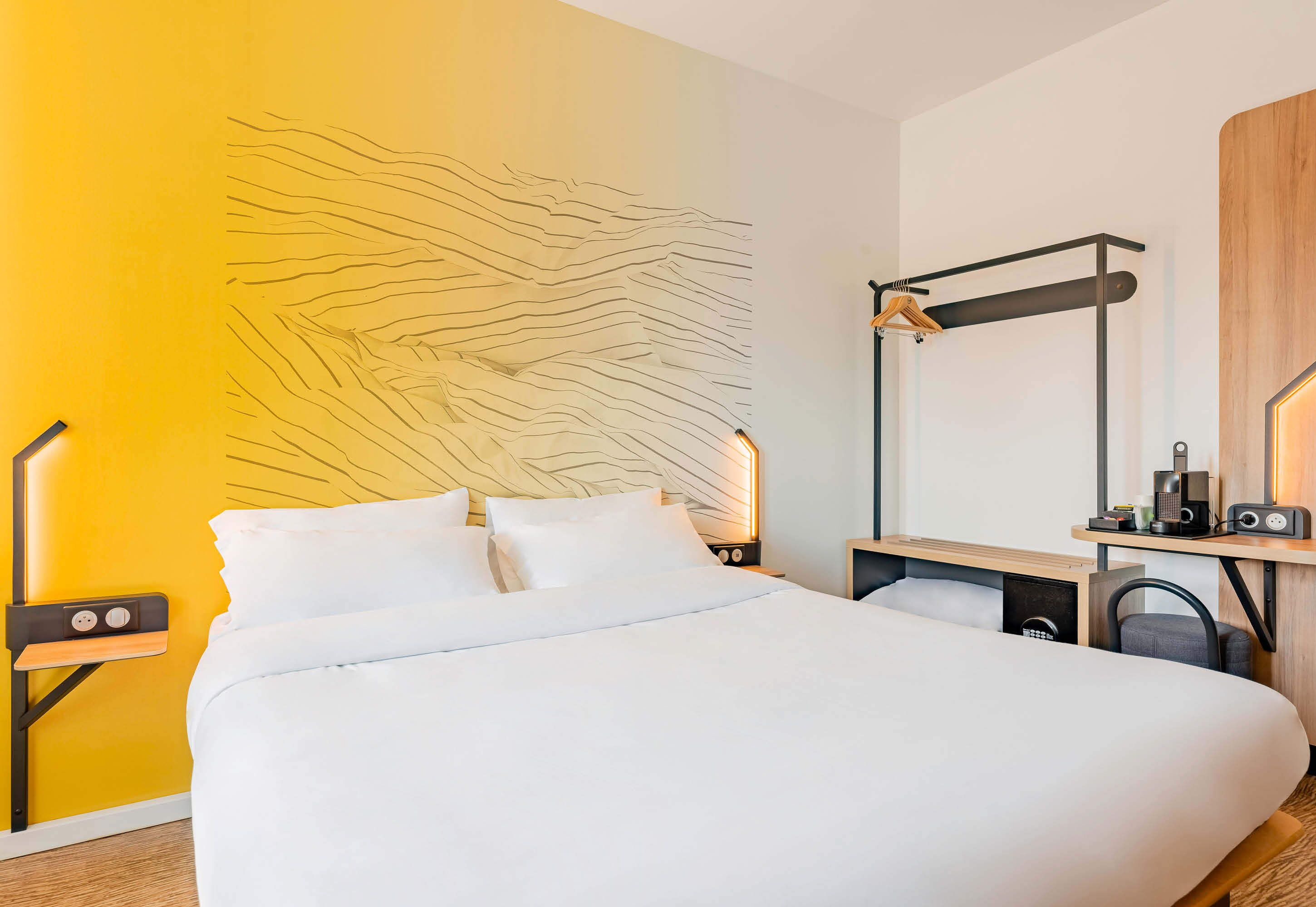 Une chambre d’hôtel moderne, équipée d’un lit, d’un bureau avec une lampe et d’un mur au design original.