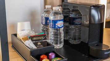 A hotel desk with a coffee maker, two bottles of water, a box of sugar, and a container of chocolates.