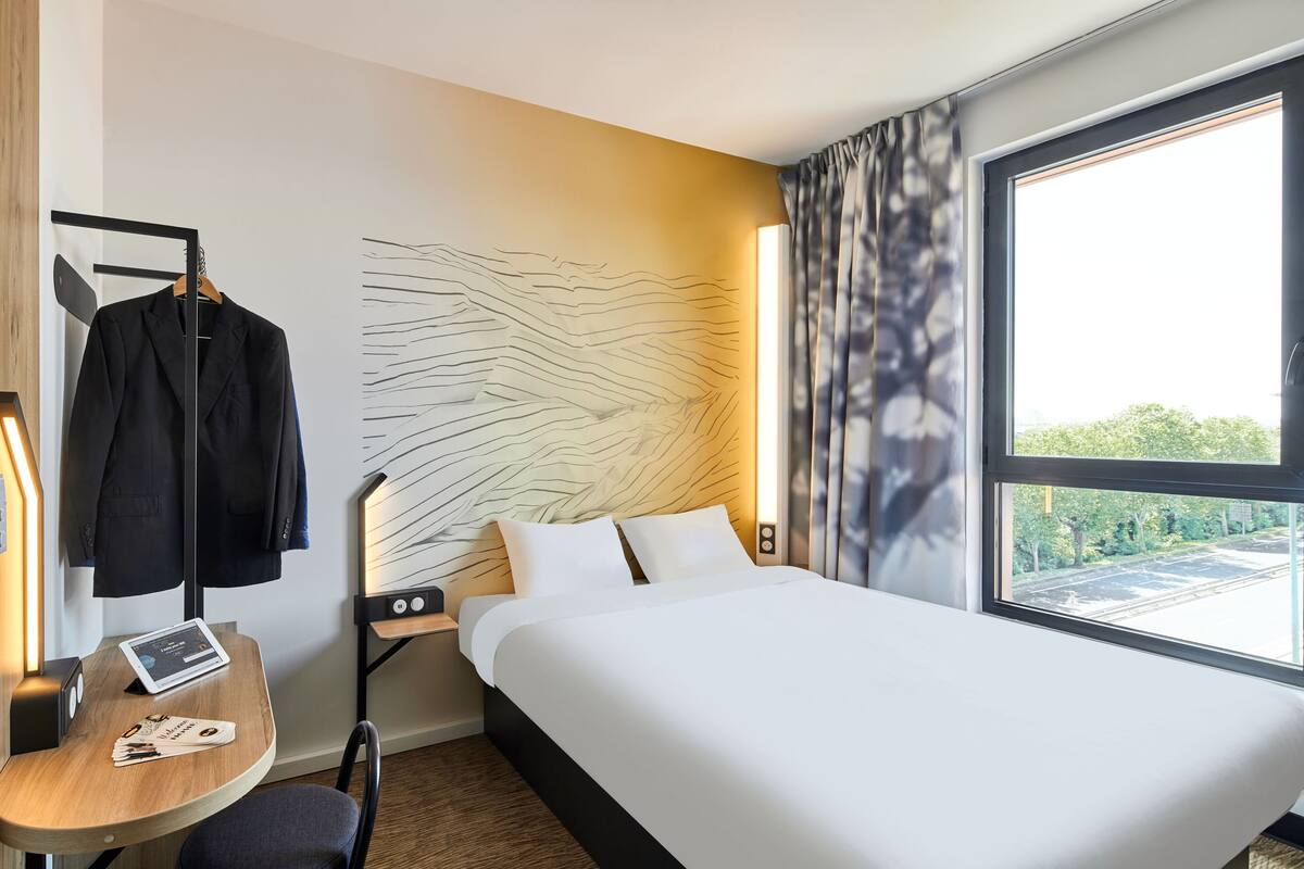 Una habitación de hotel con una cama grande, un escritorio con silla, un perchero y vistas a la calle a través de un ventanal amplio.
