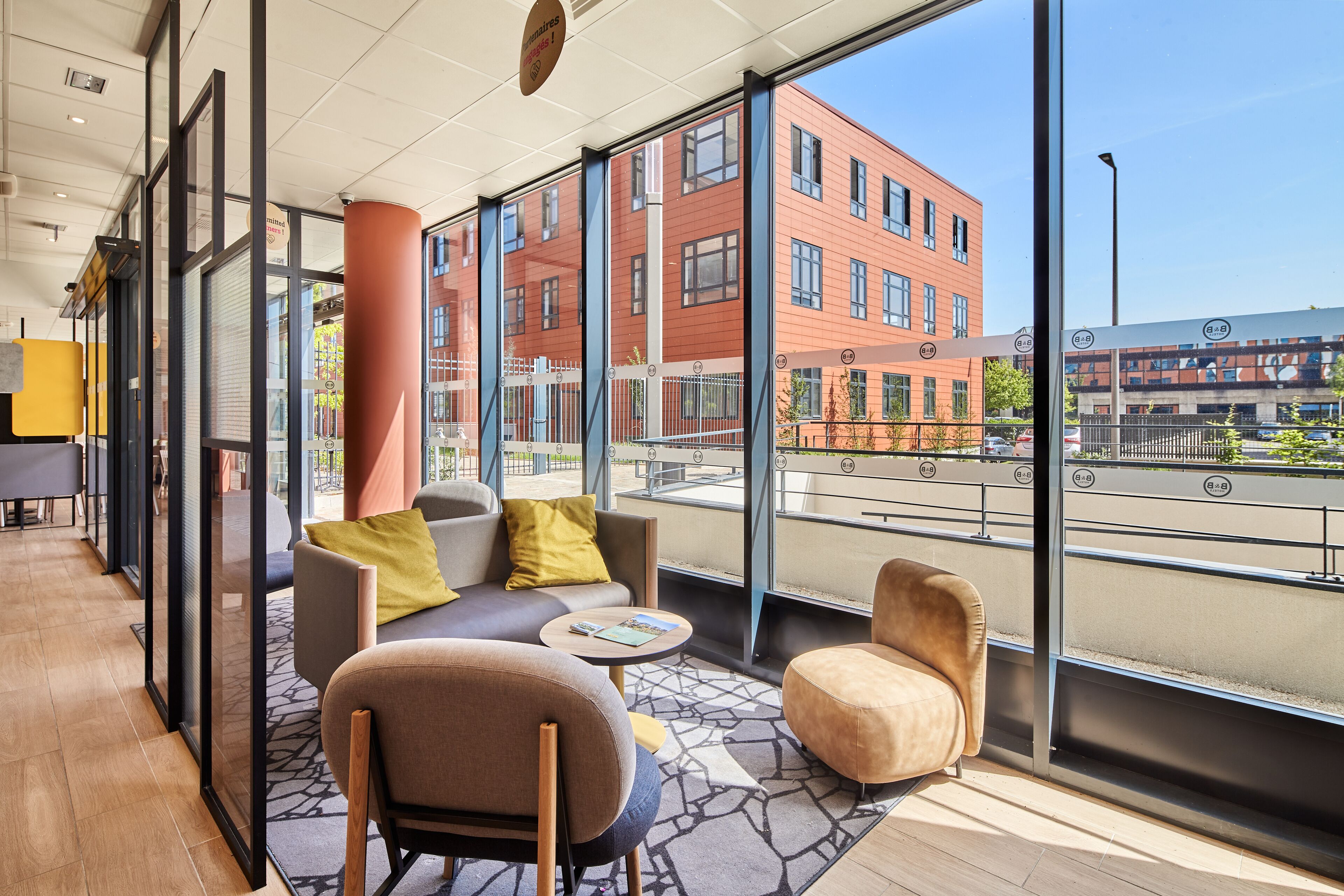 A modern hotel lobby with a seating area, large windows, and a view of a red building. - B&B HOTEL Argenteuil (Argenteuil)