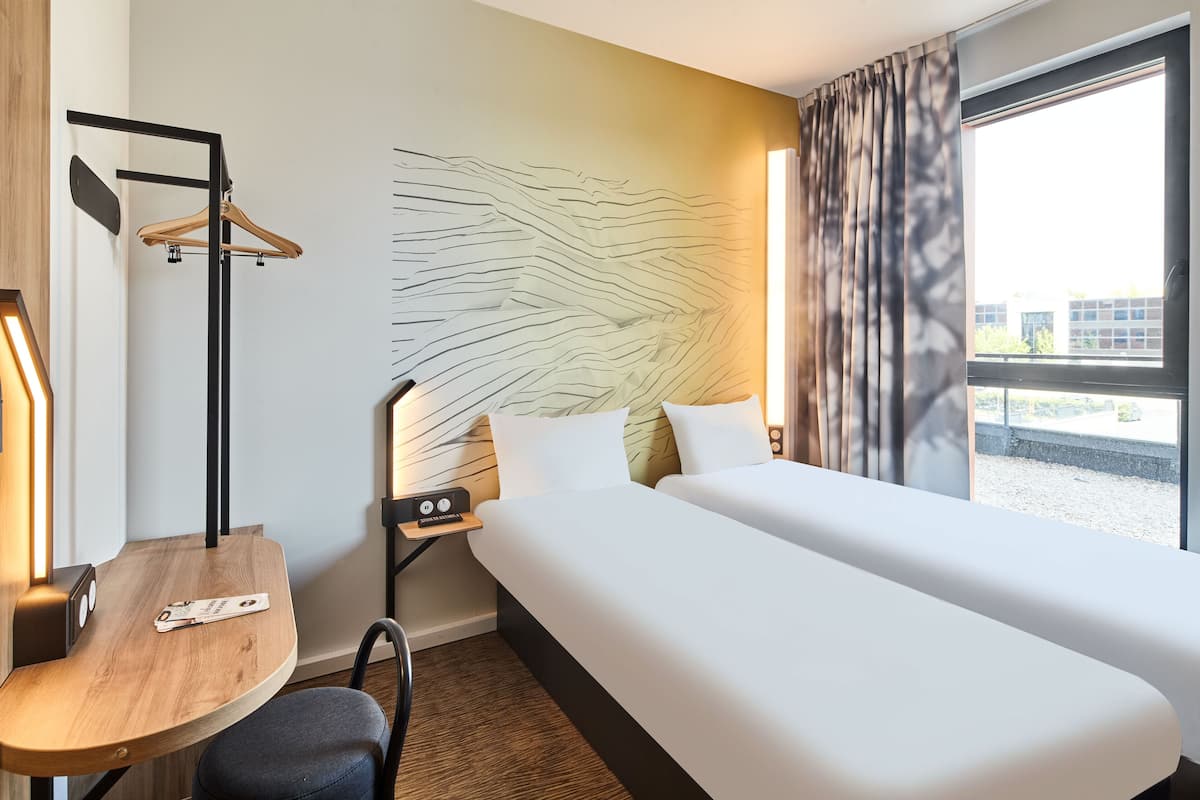 Habitación de hotel con cama, escritorio, silla, perchero y ventana con cortinas.