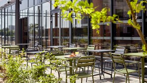 An outdoor café with tables and chairs, a glass building in the background, and greenery in the foreground. - B&B HOTEL Argenteuil (Argenteuil)