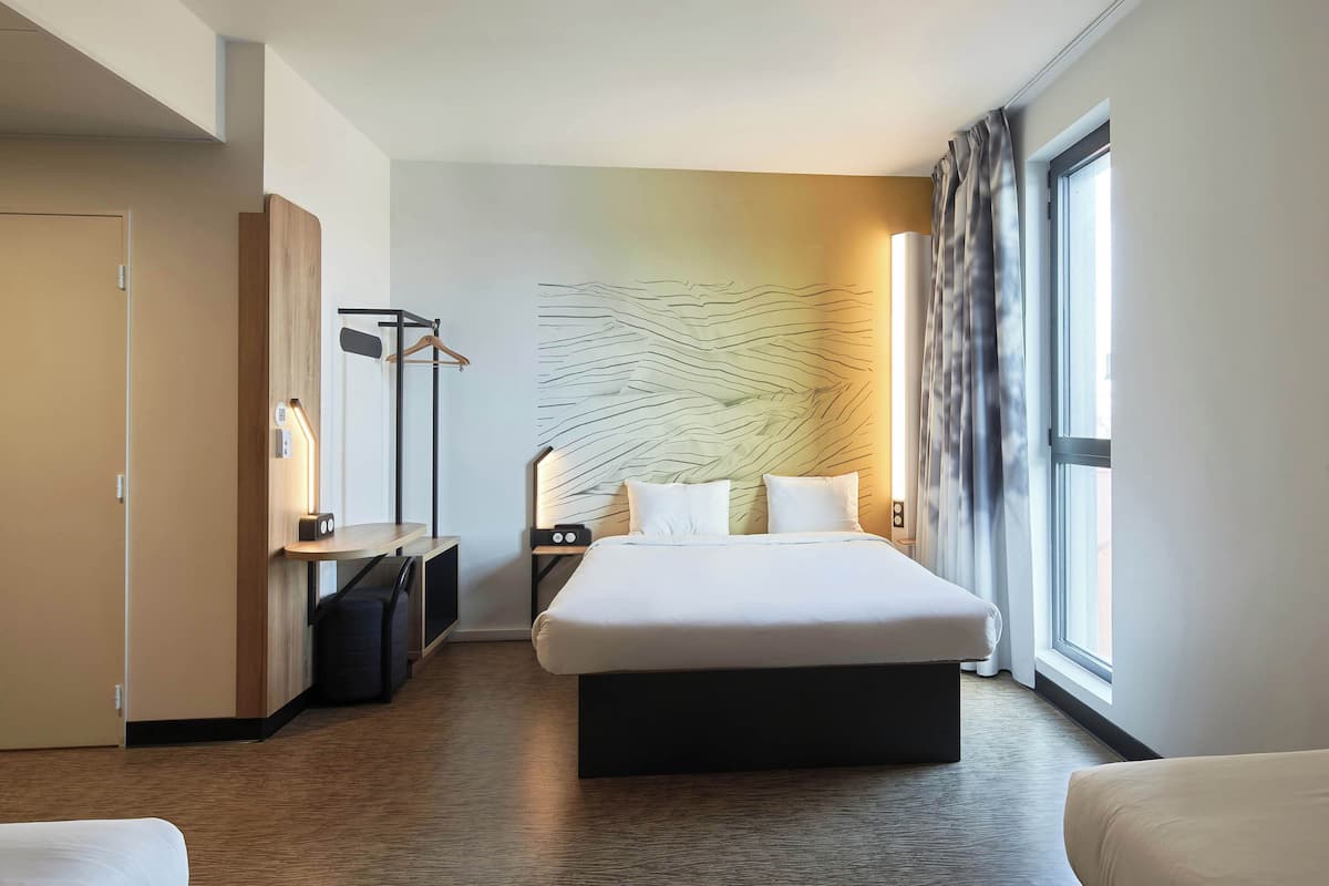 Habitación de hotel con una cama grande, un escritorio con silla, perchero y una obra de arte en la pared.