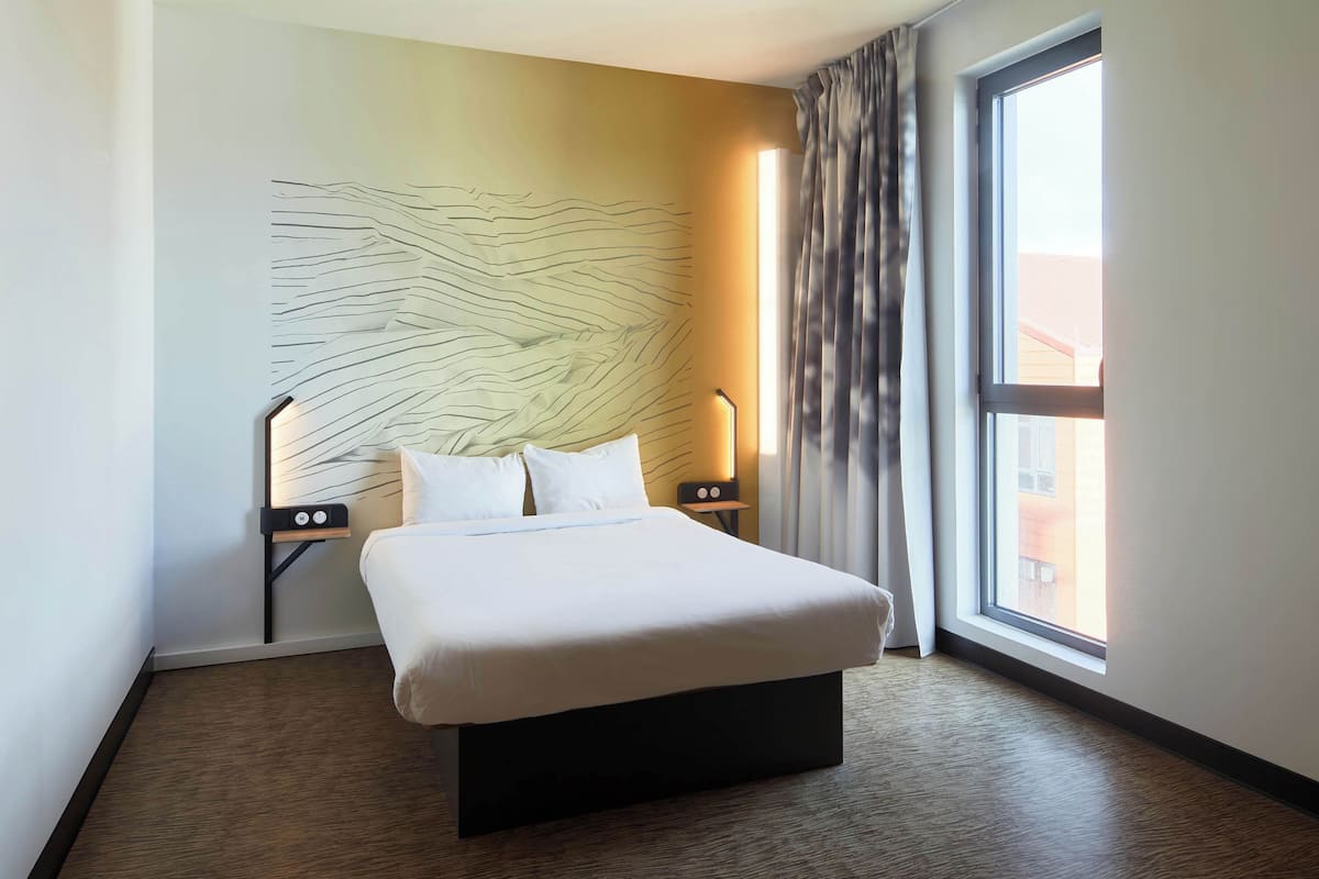 Un cuarto de hotel con una cama grande, una obra de arte moderna en la pared, una mesita de noche con lámpara y vista al exterior a través de una ventana.