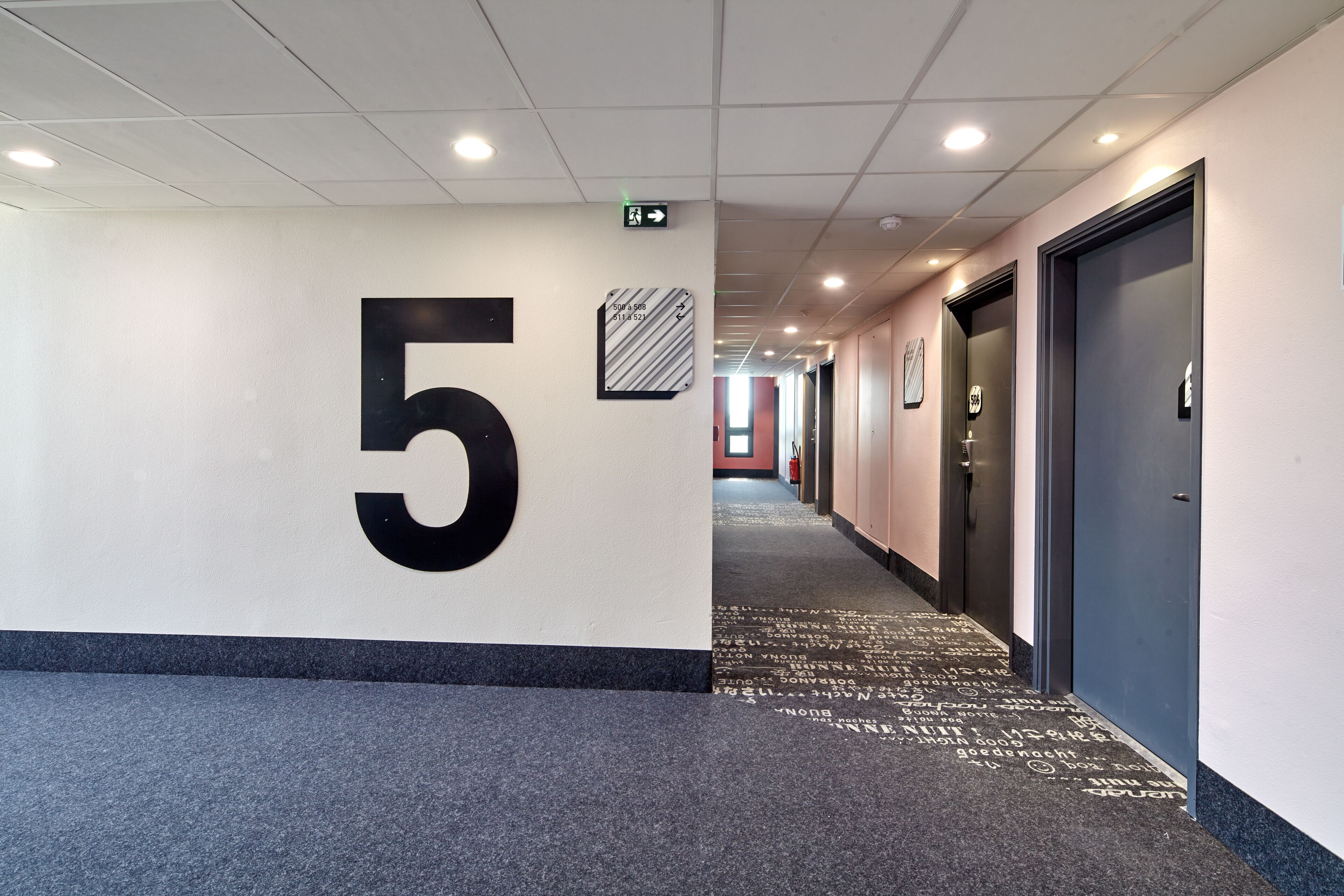 Un couloir avec un grand chiffre « 5 » peint sur le mur, indiquant le cinquième étage. Il y a des portes sur la droite et un tapis avec du texte.