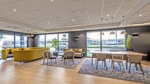 Un moderno vestíbulo de hotel con un sofá amarillo, un área de comedor con sillas de madera y grandes ventanales que dan a un estacionamiento.