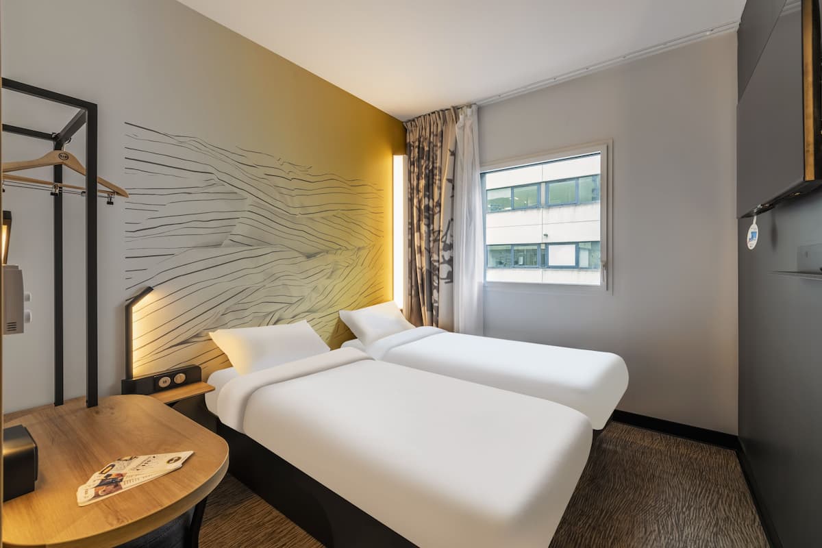 Une chambre d’hôtel avec deux lits, une table en bois, une tête de lit ornée d’un motif lumineux, une fenêtre avec des rideaux et une vue sur un bâtiment à l’extérieur.
