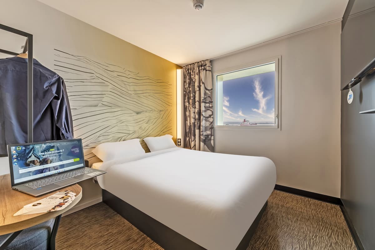 Une chambre d’hôtel avec un lit, un ordinateur portable posé sur une table en bois, une vue sur le ciel par la fenêtre et un mur orné d’un motif géométrique.