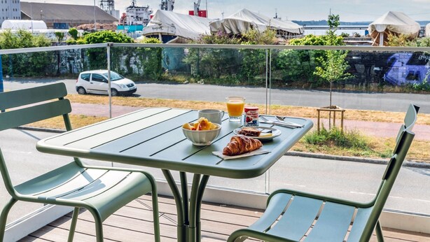 Un balcón con mesa listo para el desayuno, con vistas a un puerto deportivo con barcos y edificios industriales.