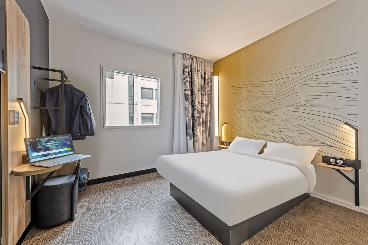 Une chambre d’hôtel moderne équipée d’un lit, d’un bureau avec un ordinateur portable, d’un porte-manteau et d’une grande fenêtre.