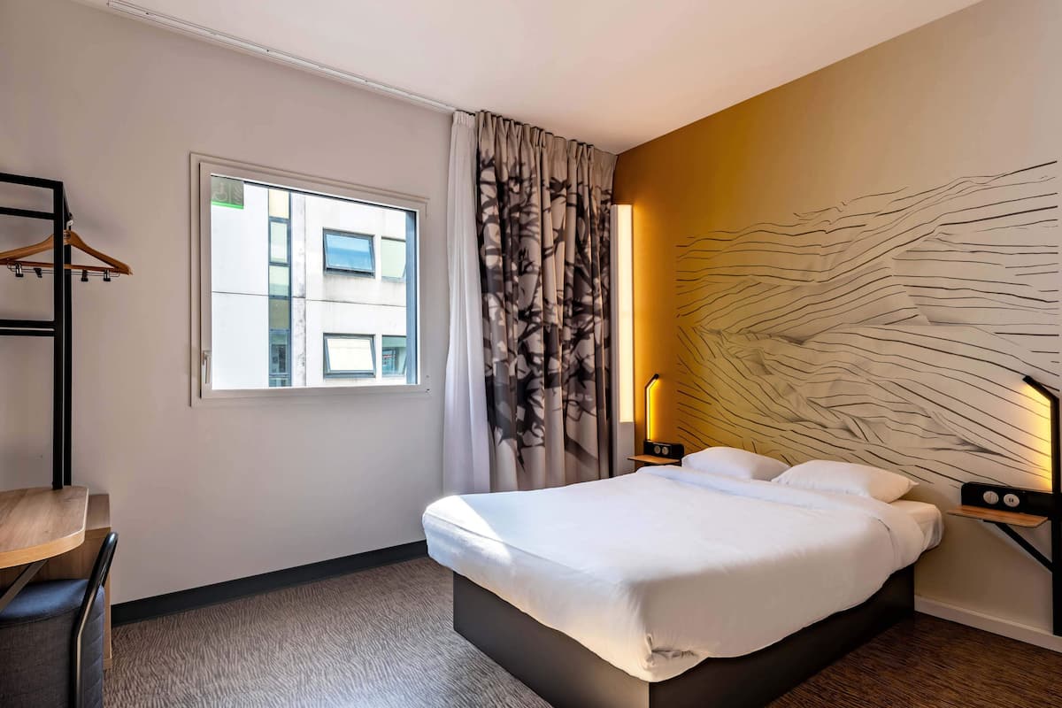 Une chambre d’hôtel comprenant un lit, un bureau, une chaise, une fenêtre avec des rideaux et un mur orné d’un luminaire original.