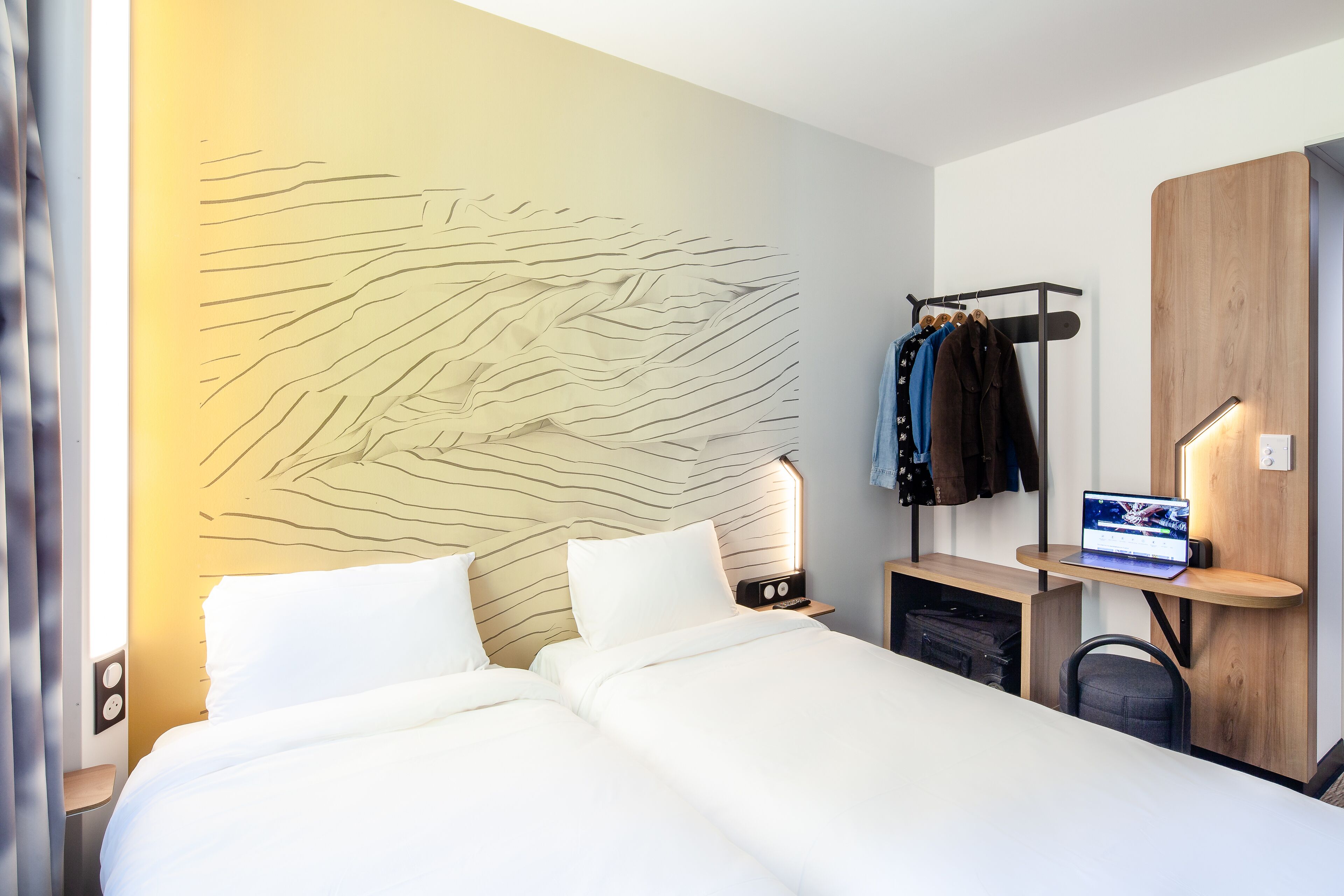 A modern hotel room with a large bed, a desk with a laptop, a coat rack, and a unique wall design.