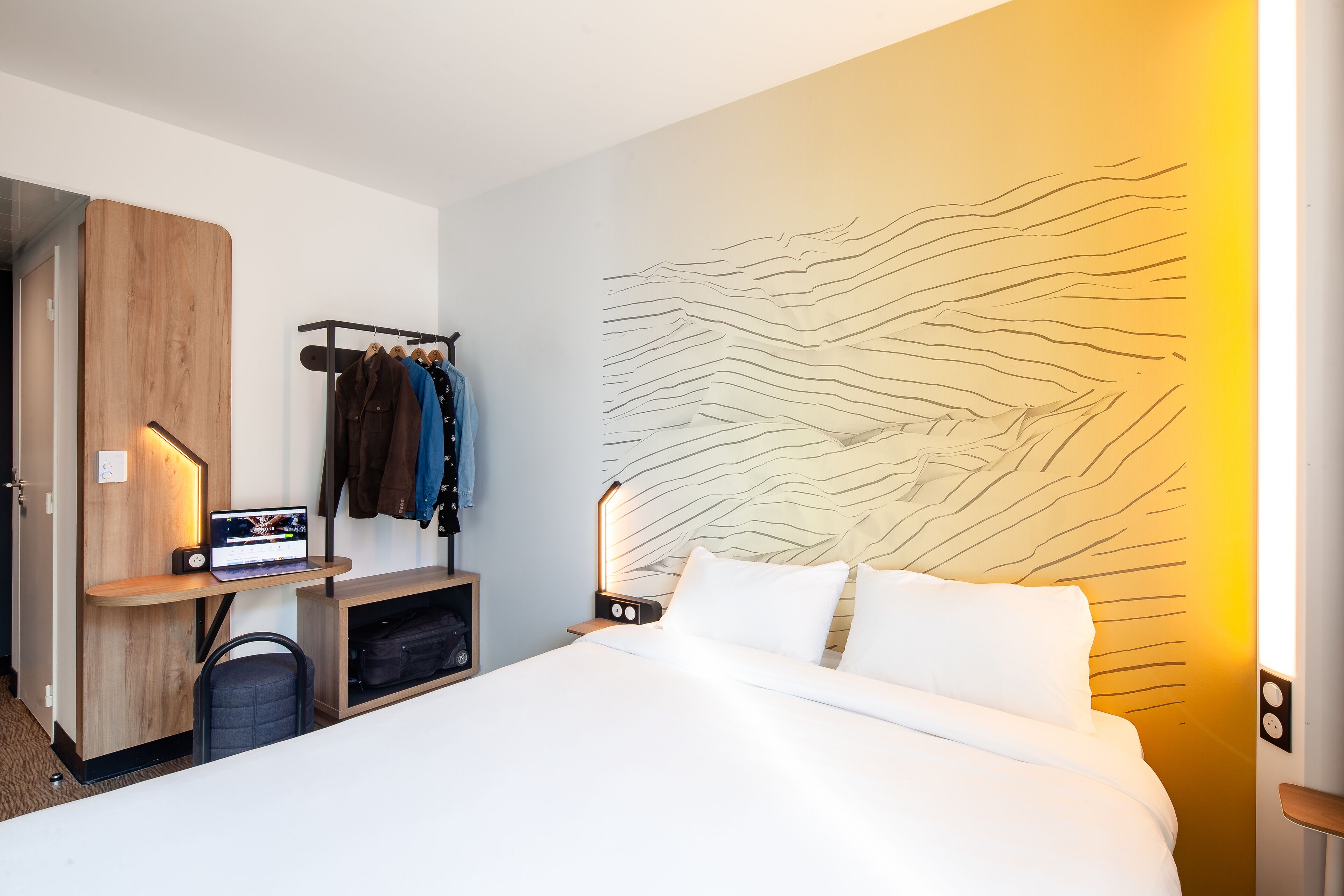 Una habitación de hotel moderna con una cama, un escritorio con una computadora portátil, un perchero con chaquetas y una pared con una textura única.