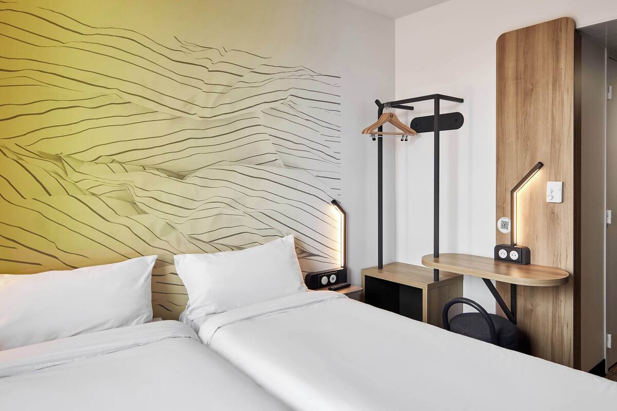 Une chambre d’hôtel moderne, équipée d’un lit, d’une table de chevet avec une lampe, d’une armoire en bois et d’un porte-manteau.