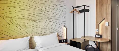 A modern hotel room with a bed, a bedside table with a lamp, a wooden wardrobe, and a coat rack.