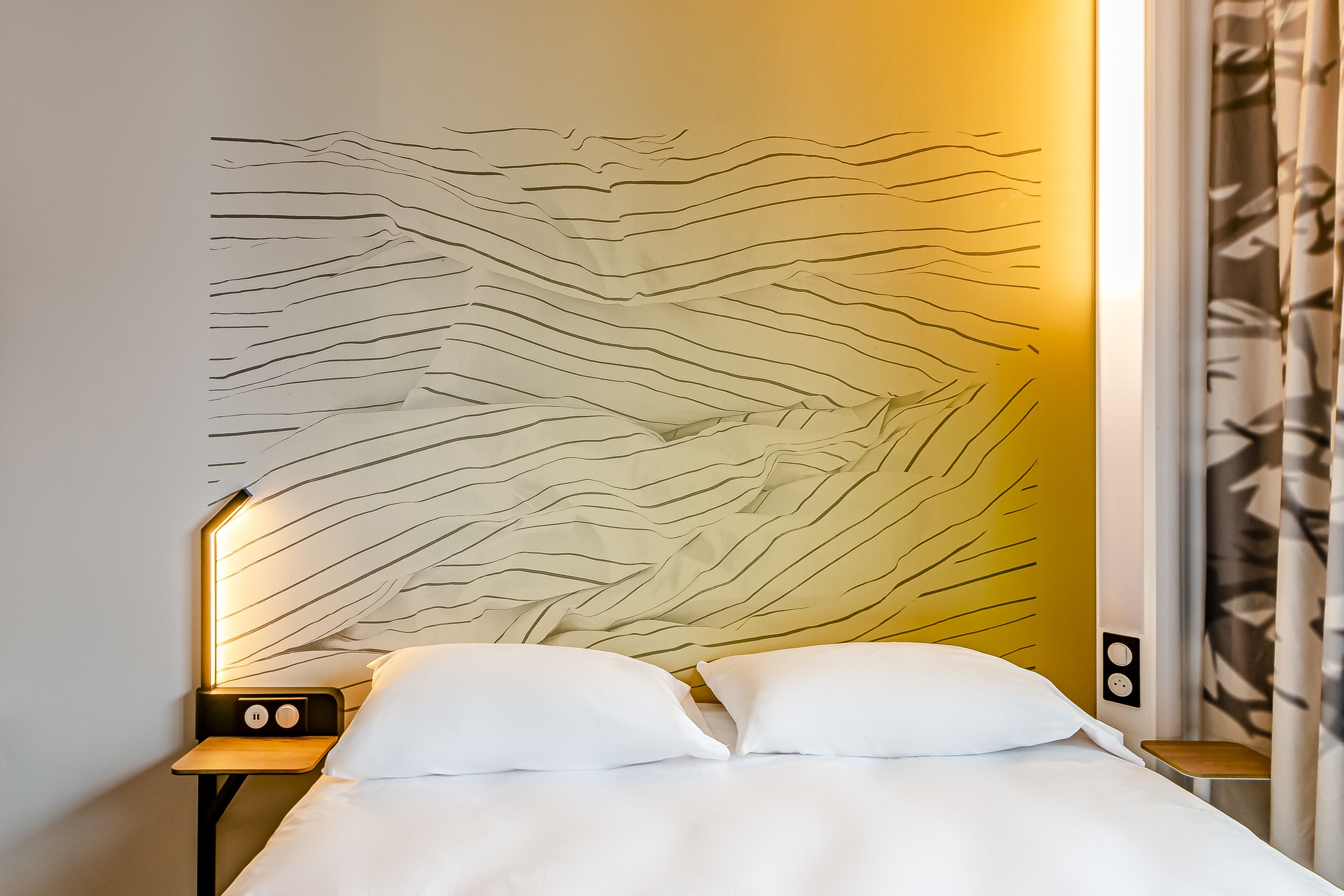 A neatly made bed with white bedding, a wall with a geometric pattern, and a nightstand with a lamp.