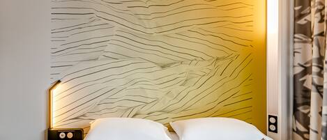 A neatly made bed with white bedding, a wall with a geometric pattern, and a nightstand with a lamp.