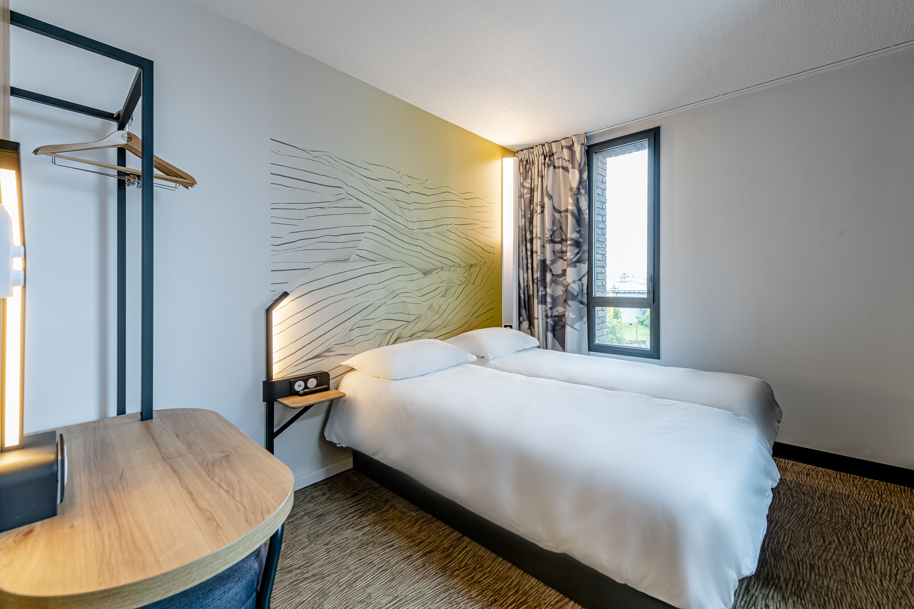 A modern hotel room with a large bed, a bedside table, a wall-mounted light, and a window with curtains.