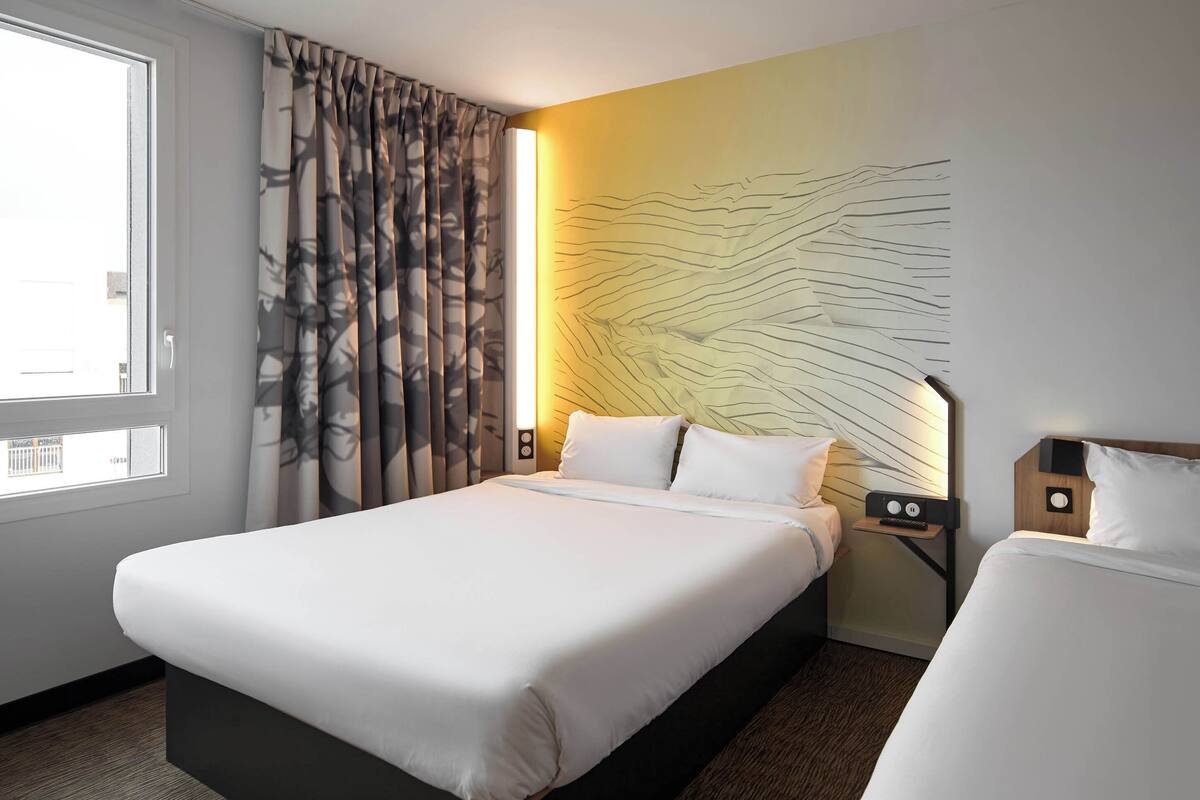 Une chambre d’hôtel avec un grand lit, une table de chevet avec une lampe, une œuvre d’art accrochée au mur et une fenêtre avec des rideaux.