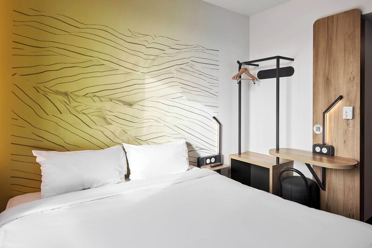 Une chambre d’hôtel moderne avec un grand lit, une tête de lit en bois, un bureau avec une chaise et un porte-manteau fixé au mur.