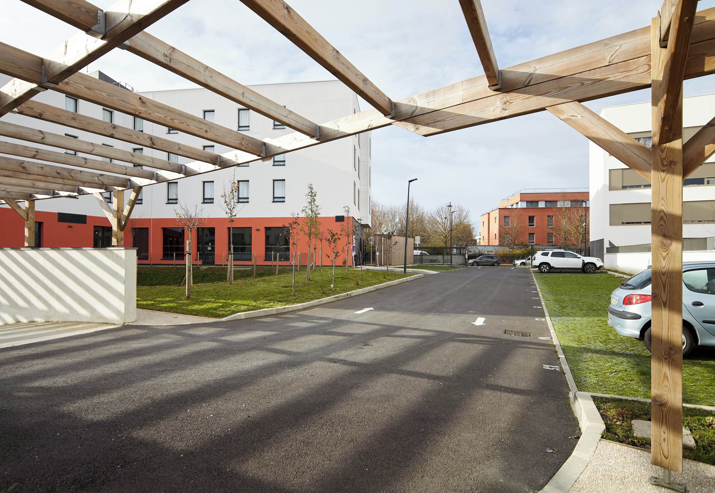 A modern building with a wooden pergola structure, a parking area, and a street with cars.
