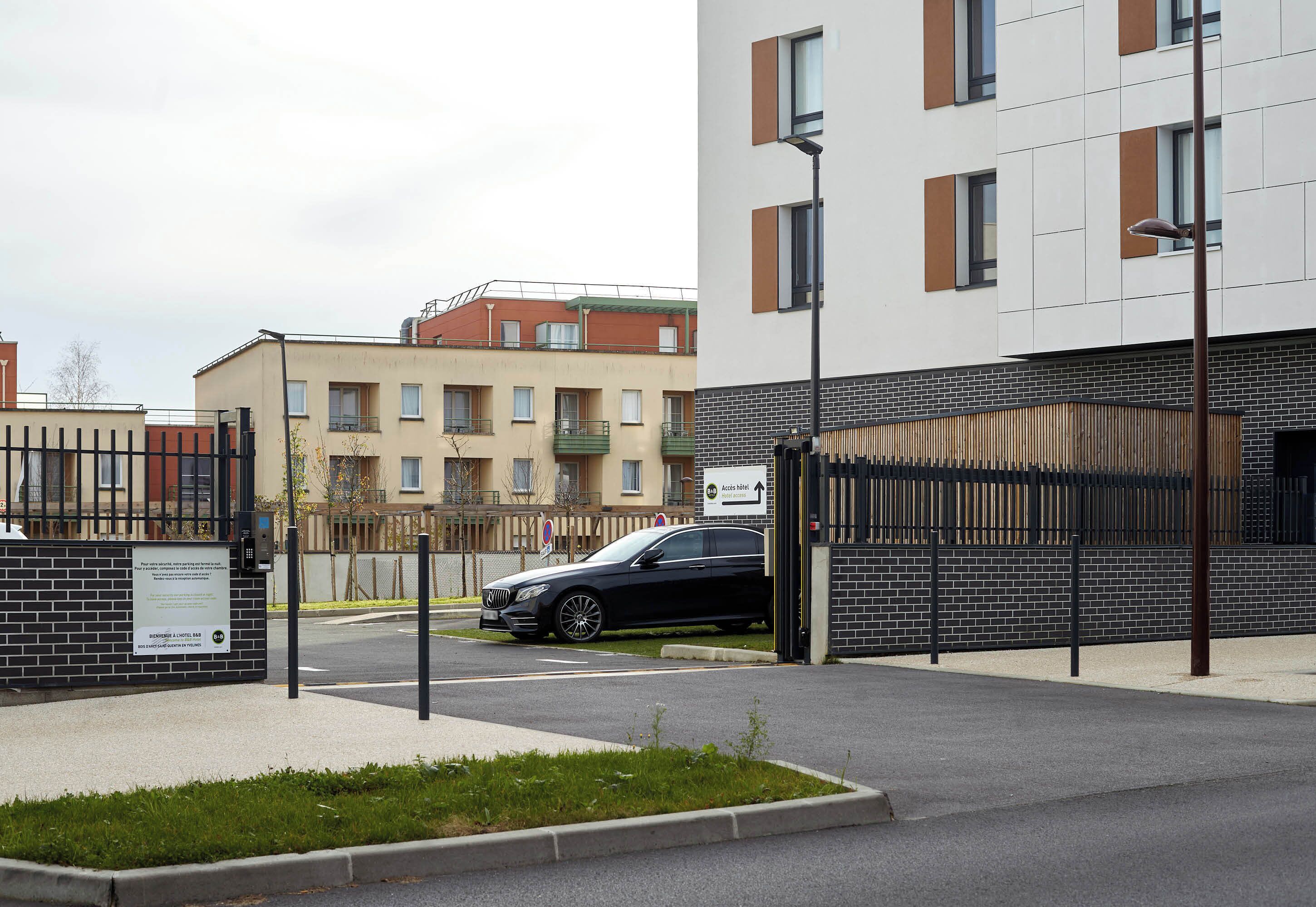 Un lotissement résidentiel fermé, avec une voiture noire garée devant un immeuble moderne à la façade en briques et un panneau indiquant une direction.