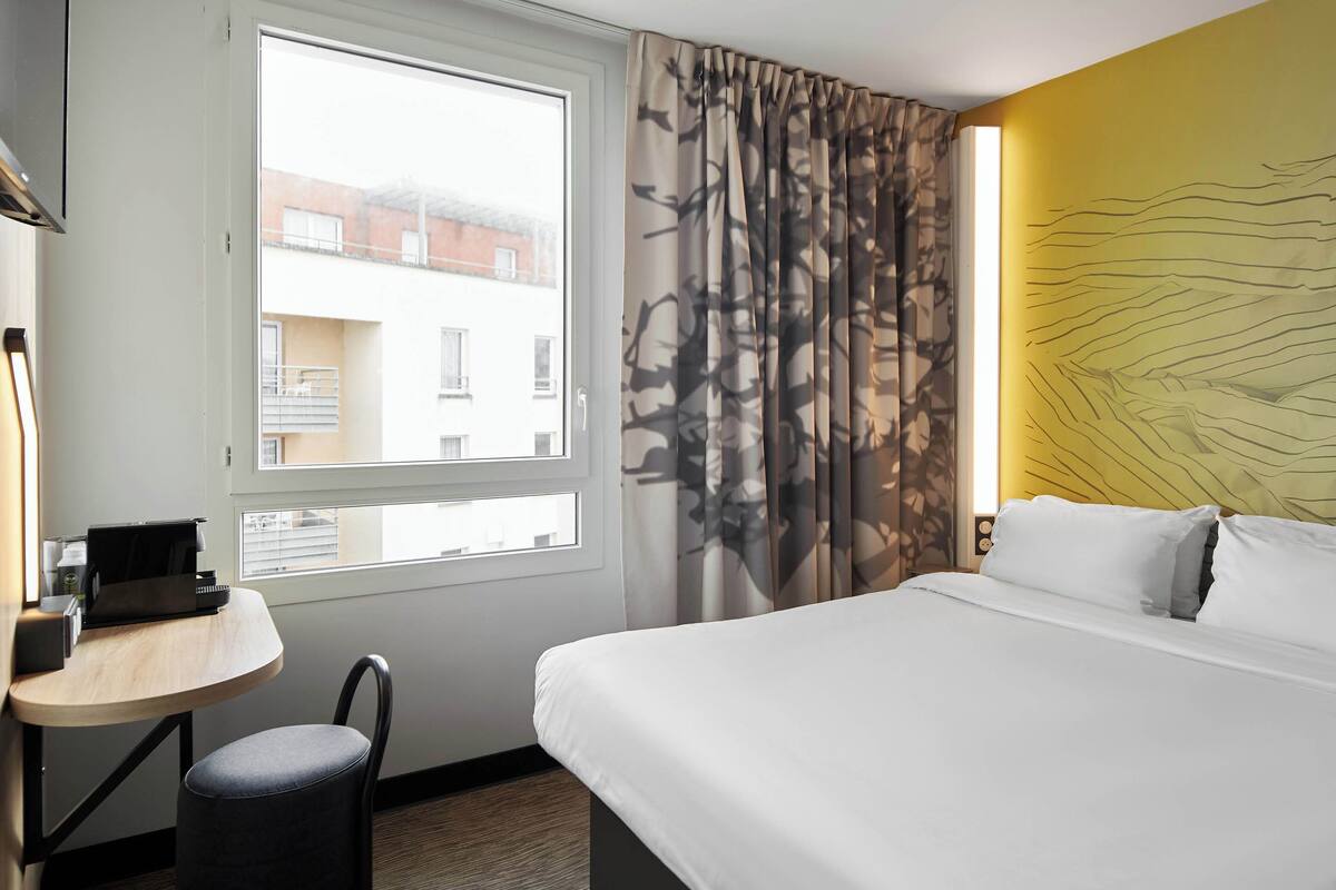 Une chambre d’hôtel avec un lit, un bureau avec un ordinateur portable, une chaise et une fenêtre avec des rideaux.