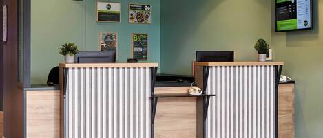 Un mostrador de recepción con encimera de madera y separadores a rayas, un televisor montado que muestra información de servicios y una planta en maceta sobre el mostrador.