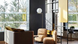 A modern living room with a sofa, armchair, and ottoman, a floor lamp, and a view through large windows. - B&B HOTEL Bois D'Arcy Saint Quentin en Yvelines (Bois-d'Arcy)