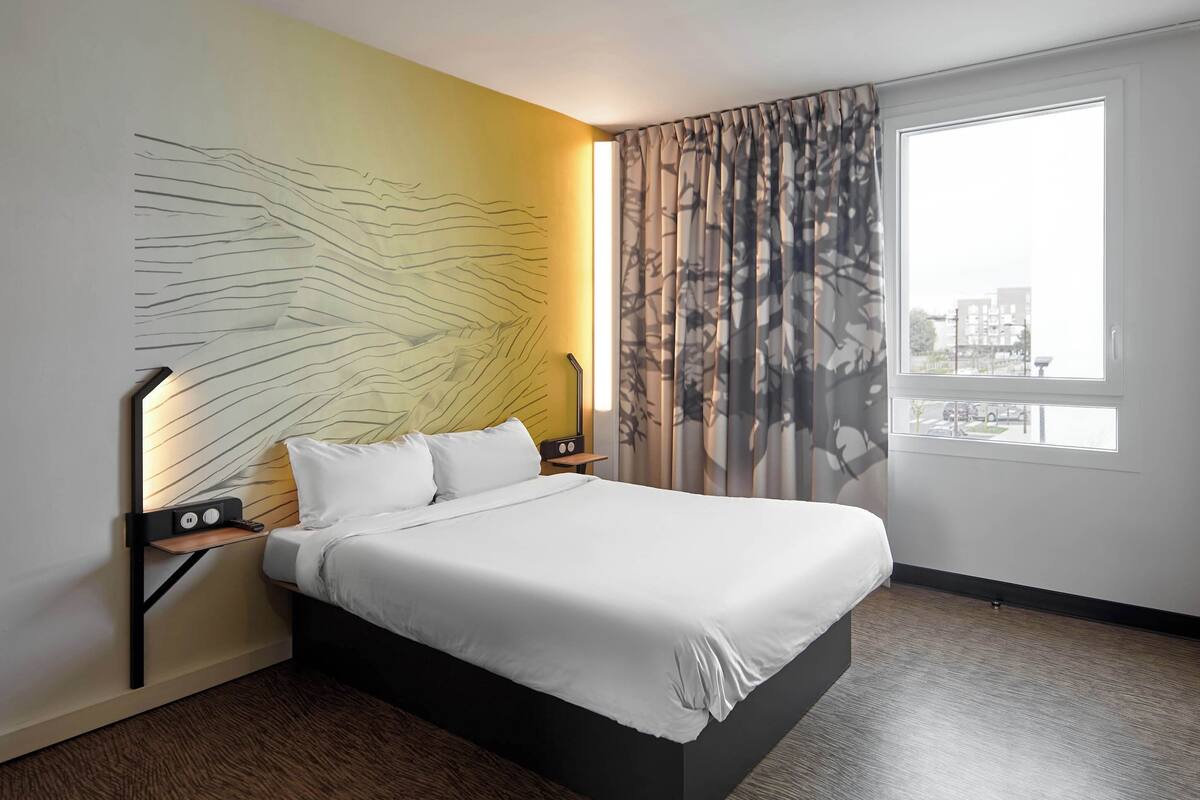 Une chambre d’hôtel moderne avec un grand lit, un luminaire fixé au mur, une fenêtre avec des rideaux et une vue sur la ville.