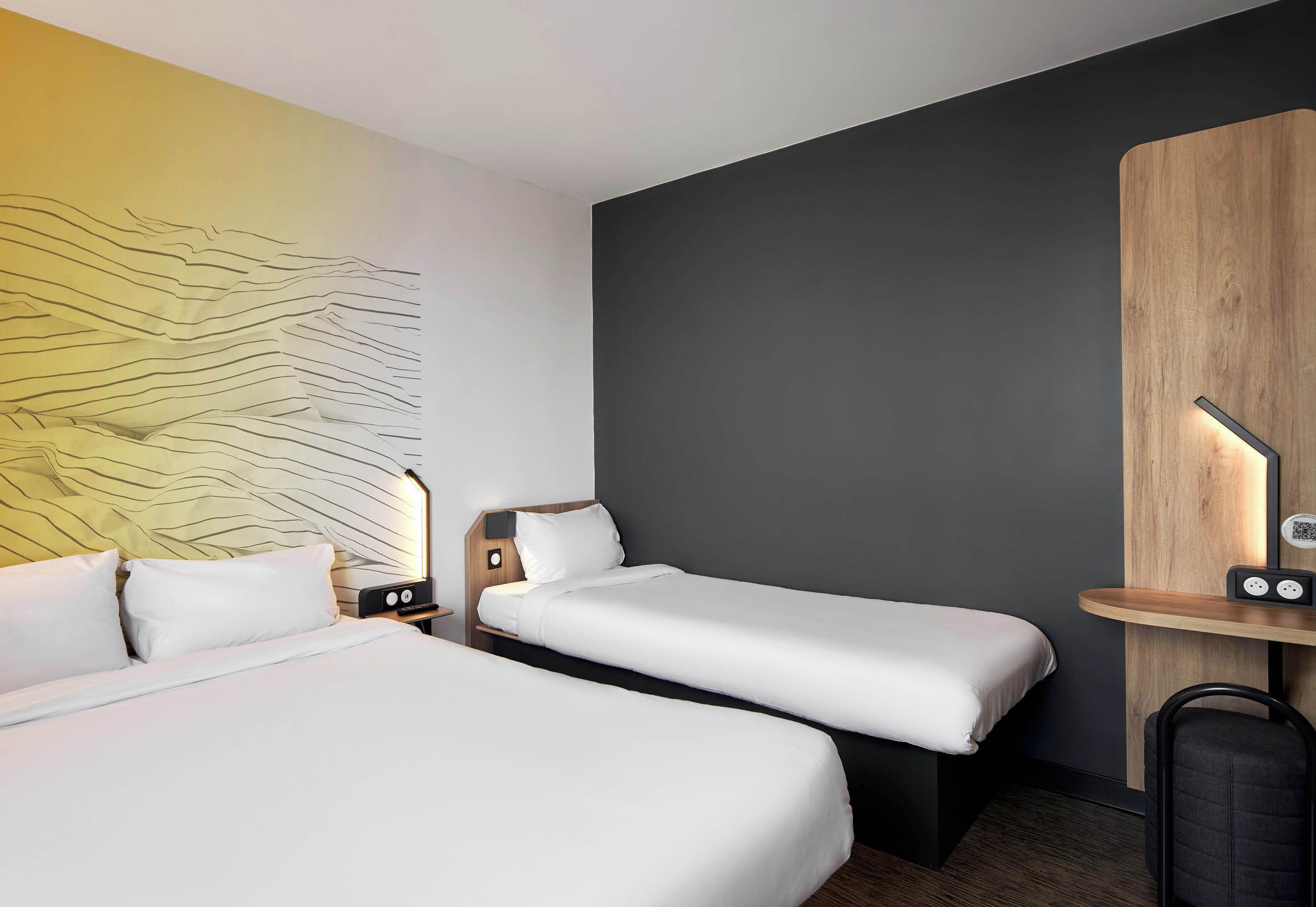 Ein Hotelzimmer mit zwei Betten, einem hölzernen Kopfteil, einem Schreibtisch mit Lampe sowie einer Wand mit strukturierter Gestaltung.
