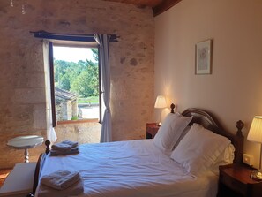Ein Schlafzimmer mit einem Bett, zwei Nachttischlampen, einem Nachttisch, einem Fenster mit Vorhängen und Blick ins Grüne.