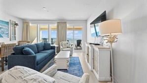 Ein modernes Wohnzimmer mit blauem Sofa, weißem Sessel und einem großen Flachbildfernseher. Durch große Fenster ist der Blick aufs Meer möglich.