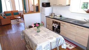 Eine kompakte Küche mit Kühlschrank, Backofen und Spüle. Ein Esstisch mit gemustertem Tischläufer und Stühlen lädt zum Verweilen ein. Im Wohnbereich befinden sich eine Couch und ein kleiner Tisch mit einem Blumenvasen.