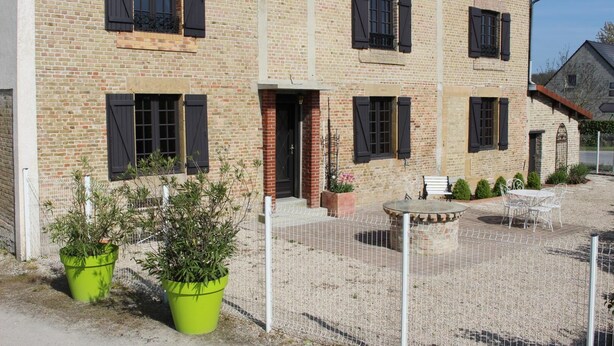 A two-story brick building with black shutters on the windows, a small outdoor seating area with a table and chairs, and potted plants. - 1843 LE RUISSEAU DU MOULIN (SEUIL)
