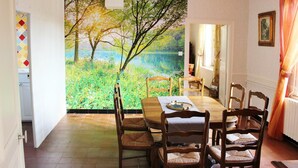 A dining area with a large painting of a forest and lake, wooden chairs, a table with a placemat, and a chandelier. - 1843 LE RUISSEAU DU MOULIN (SEUIL)