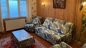Una sala de estar acogedora con un muro de madera, un sofá estampado, dos sillones, una mesa de centro y un ventanal con cortinas.