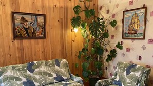 Una habitación con paredes de madera, una planta grande en maceta, dos sofás con motivos de hojas tropicales y cuadros de temática marina enmarcados.
