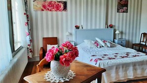 Ein Schlafzimmer mit einem Bett, einem Nachttisch, einem Sessel, einem Tisch mit Blumenvase und einem fenster mit Vorhängen.