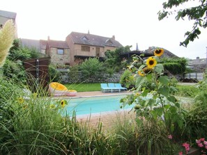 A pool area with a clear view of the pool, surrounded by lush greenery and a house in the background. - LE Grain D'amour - Chooz (CHOOZ)