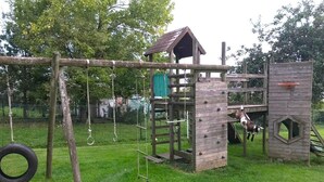 A wooden play structure with a climbing wall, a hanging tire swing, and a goat enclosure.